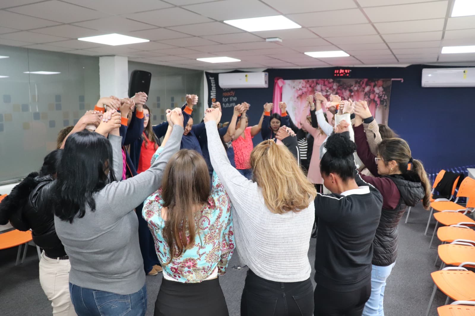 Grupo numeroso de participantes com os braços erguidos durante oficina em sala de empresa